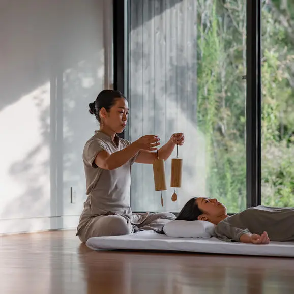 Wellness practitioner performs a massage ritual on a guest at Amanemu resort, sunlit room with large windows overlooking greenery.