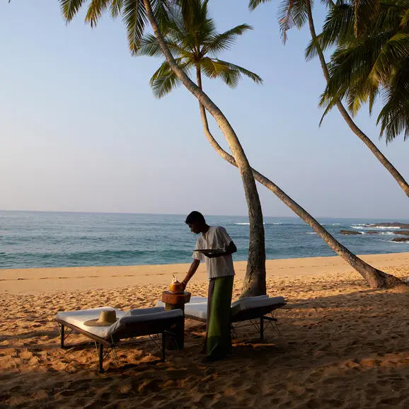 A guest relaxes in a wooden chair beneath palm fronds on Amanwella's sandy beach, with calm waters and coastline beyond.