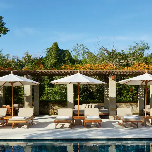 Poolside lounge area at Aman Villas at Nusa Dua with pergolas, seating, and mountain views.