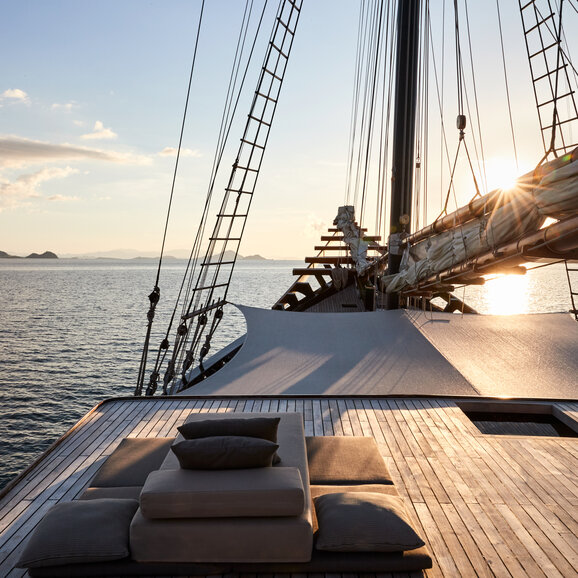 Wooden deck of a sailing vessel at Amandira with rigging and cushioned seating, overlooking calm waters at sunset.