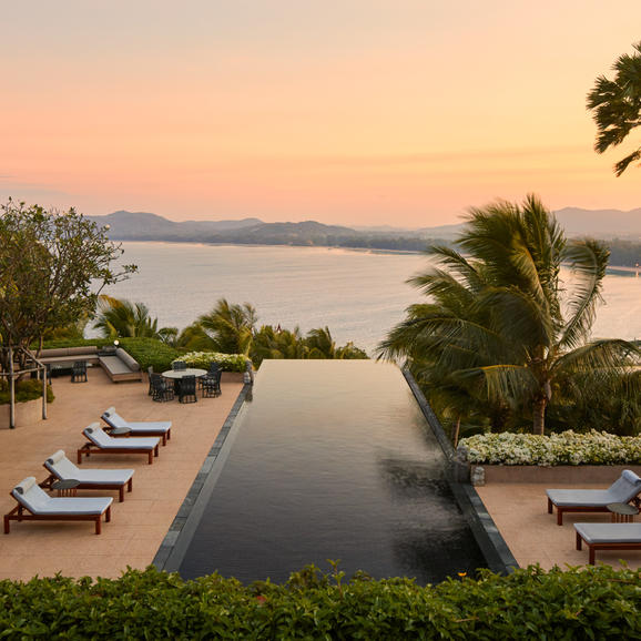 Amanpuri villa with infinity pool overlooking the Andaman Sea at sunset, flanked by palm trees and sun loungers.