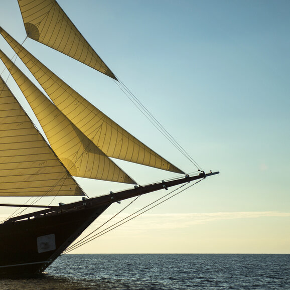 Amandira's prow detail with billowed sail against a clear sky and sea horizon.