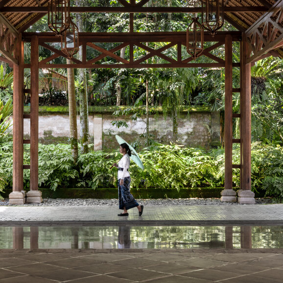 安缦黛宁度假村大堂内，一位访客站在木制走廊旁观景，绿植环绕，水池倒映周围景致。
