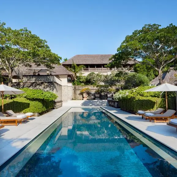 Rectangular swimming pool at Aman Villas at Nusa Dua, flanked by tropical gardens and lounge areas under clear sky.