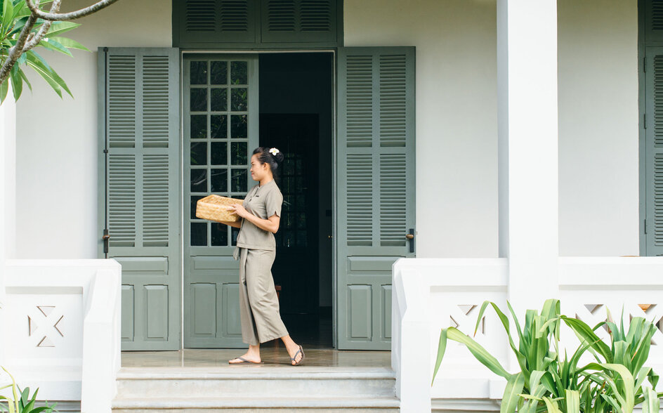 A woman stands on the stone terrace of an Amantaka suite, framed by white columns and shuttered doors, with lush greenery flanking the entrance.