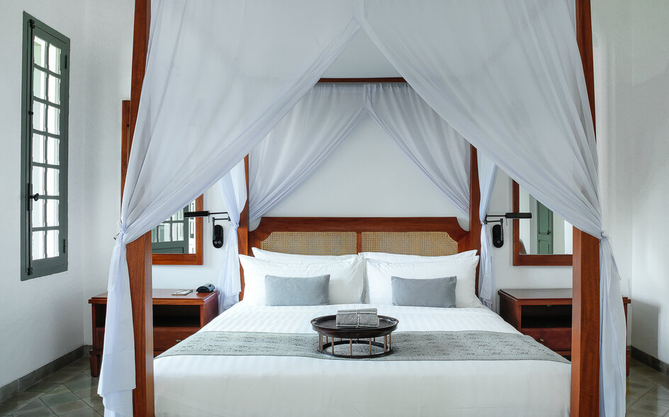 Khan Suite principal bedroom at Amantaka, featuring a four-poster bed with white drapes and wooden bedside tables.