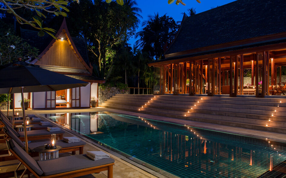 Evening view of an illuminated swimming pool and pavilion at Amanpuri, surrounded by tropical trees under a twilight sky.