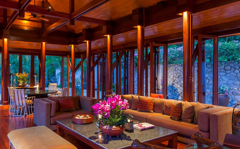 Amanpuri's dining pavilion with wooden beams, floor-to-ceiling windows overlooking the sea, and a long table set for dinner at dusk.