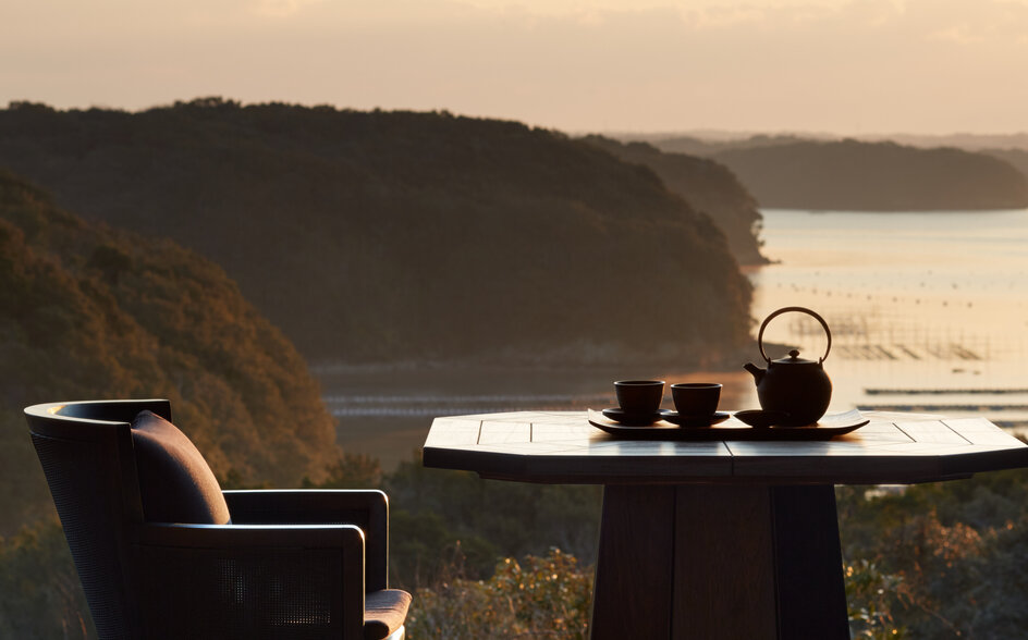 Dining table with chair overlooking forested coastline at Amanemu at sunset.