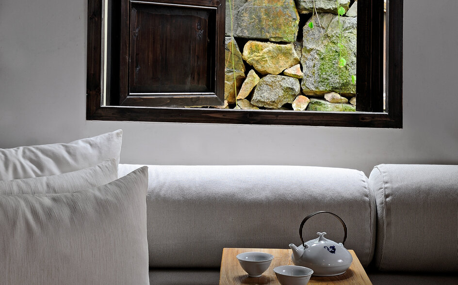 Amanfayun villa interior with grey sofa, white pillows, and wooden table framed by window overlooking stone courtyard.