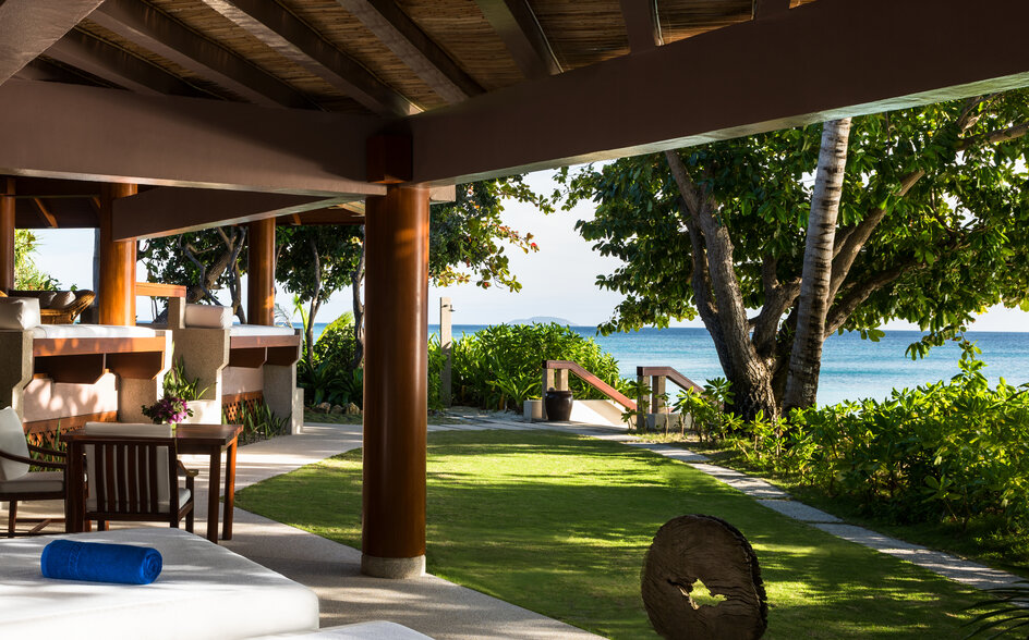 Amanpulo resort one-bedroom villa with covered terrace, wooden beams, and views across manicured gardens towards the sea.