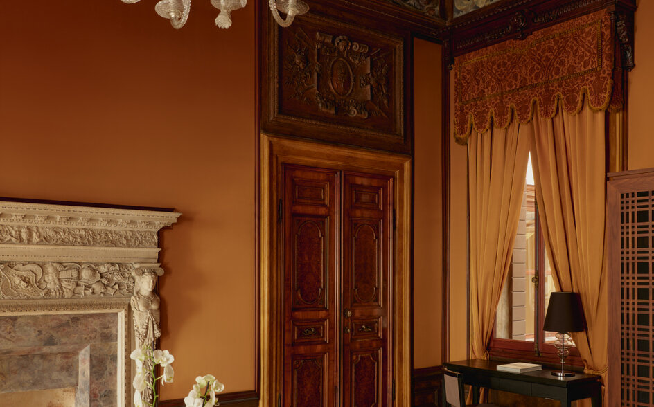 Living area at Aman Venice with ornate fireplace, chandelier, and period furnishings in warm terracotta tones.