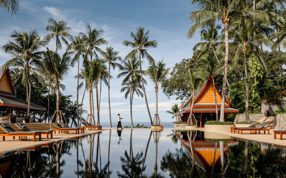 Amanpuri's wooden pavilions reflect in still water surrounded by palm trees at dawn.