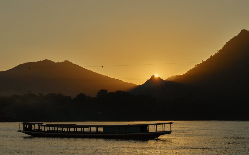 Wooden boat silhouetted on the Mekong River at sunset, mountains rising beyond at Amantaka.