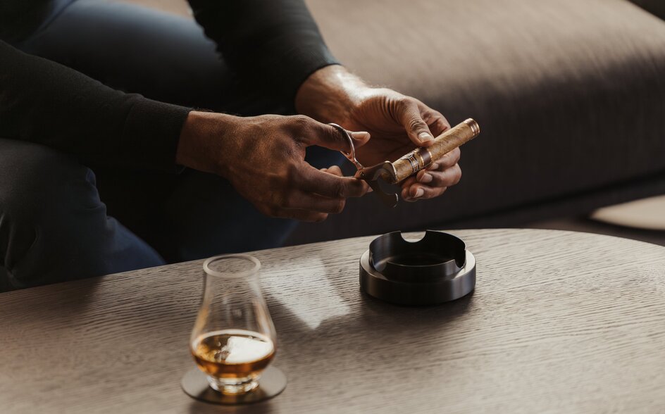 Hand holding cigar over ashtray at Aman Rosa Alpina's cigar lounge in Italy.