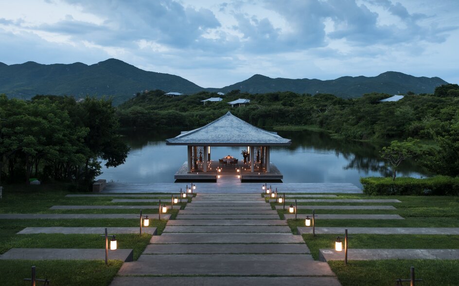 Wooden dining pavilion lit by lanterns on a lake at Amanoi, Vietnam, at dusk with forested mountains beyond.