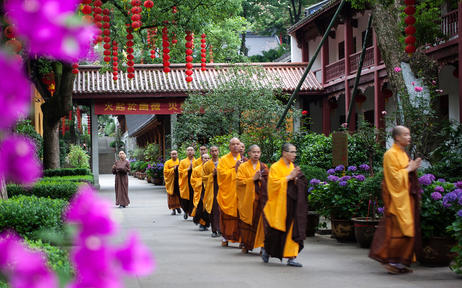 Buddhist monks in saffron robes walking through Amanfayun's traditional Chinese village courtyard, framed by magenta flowers.