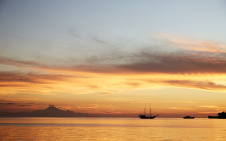 Sunset over calm waters at Amandira, with a sailing vessel silhouetted against golden and orange skies.