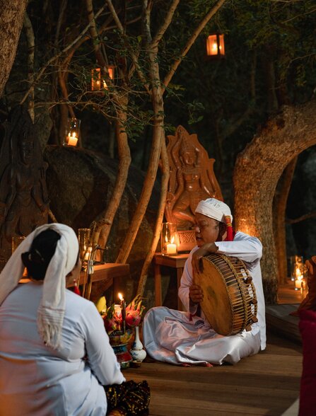 Two guests in white clothing sit beneath ancient trees with hanging lanterns at Amanoi, Vietnam.