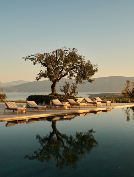 Amanzoe's main pool at sunset, with a solitary tree reflected in still water and loungers along the poolside.