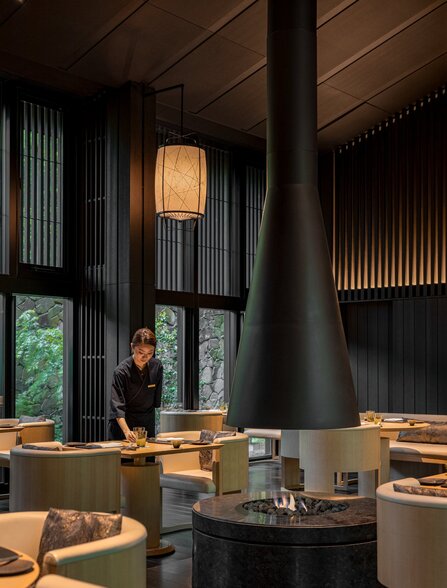 The Living Pavilion dining venue at Aman Kyoto, featuring wooden interiors, a central stone fireplace, and floor-to-ceiling windows overlooking gardens.