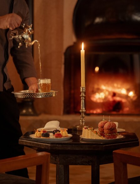 The bar at Amanjena, with a fireplace glowing warmly in the background and drinks displayed on a dark table.