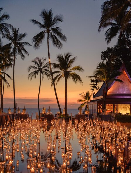 Candlelit dinner gathering at Amanpuri as sun sets behind palm trees, Thailand.