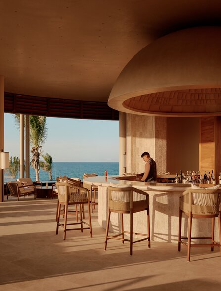 Amanvari lounge bar with curved wooden architecture, bar seating, and sea views at sunset.