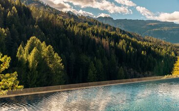 Outdoor pool at Aman Rosa Alpina with Alpine mountains and forested slopes reflected in still water.