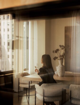 Woman seated in a light-filled penthouse living area at Aman New York, with floor-to-ceiling windows and modern furnishings.