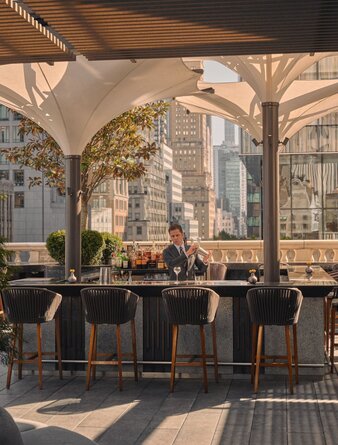 Bartender crafting cocktails at Aman New York's corner bar and garden terrace, shaded by canvas awnings.