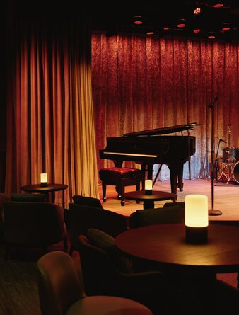Grand piano on stage at Aman New York's jazz club, framed by warm red velvet curtains.