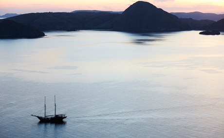 Sailing vessel anchored in calm waters at sunset, Amandira, Indonesia, with volcanic island silhouetted against twilight sky.