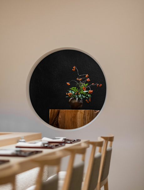 A circular black mirror with wooden frame reflects green foliage, mounted above a wooden dining table at Aman Tokyo.