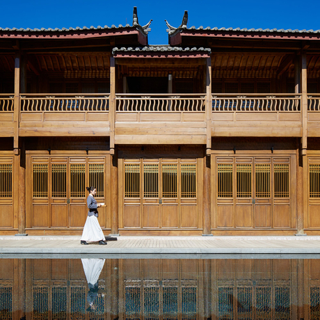 Woman in white dress seated before traditional wooden architecture at Amandayan, reflected in still water.