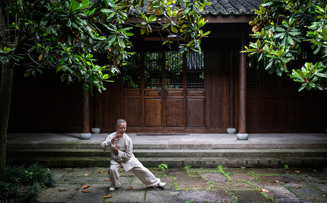 Tai chi practitioner in white clothing performing a stance in the courtyard at Amanfayun, with traditional wooden doors and green vines framing the scene.