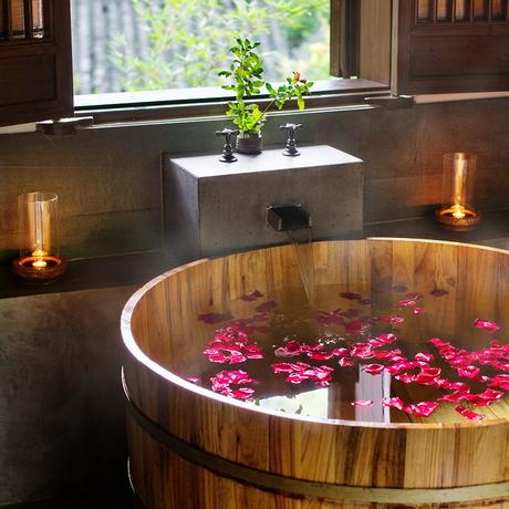 Wooden soaking tub filled with rose petals and candles at Amanfayun, with potted plant and window beyond.