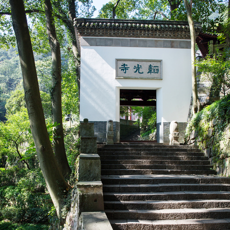 A whitewashed pavilion with traditional Chinese architecture nestled among trees at Amanfayun, featuring dark wooden steps leading to an entrance beneath overhanging branches.