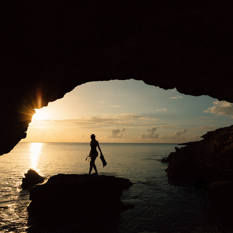 A figure walks across a rock formation within a cave opening at Amanyara, framed by golden sunset light over calm waters.