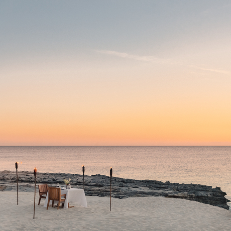 Rock formation extending into calm turquoise waters at Amanyara at sunset, with two chairs and a table on the sandy beach.