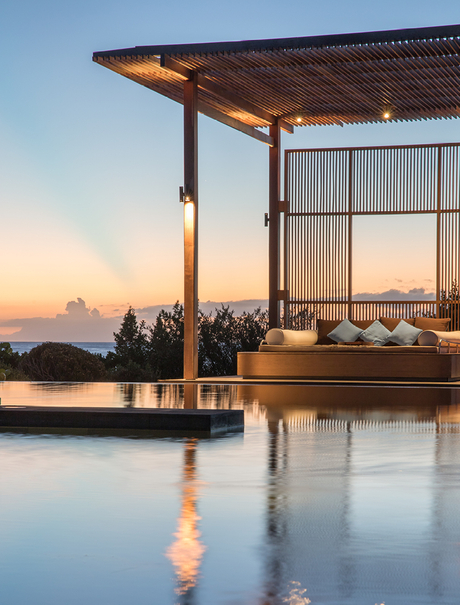 Waterfront pavilion at Amanyara with wooden pergola and floor-to-ceiling screens at sunset, reflected in still water.