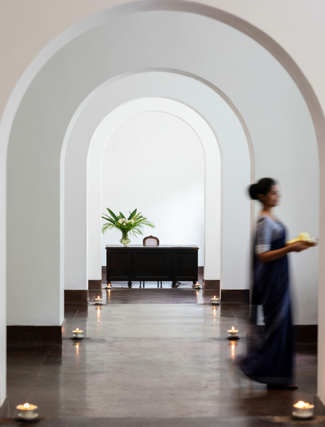 Arched corridor at Amangalla with polished grey stone floor, white walls, and a figure walking past a potted plant on a dark console table.