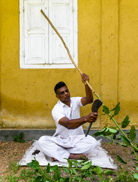 A man in white clothing sits against a golden-yellow wall at Amangalla, holding a long wooden staff upright.