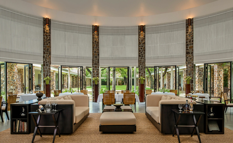 Amansara's curved reception hall with floor-to-ceiling windows overlooking gardens, featuring dark timber columns and contemporary furnishings.