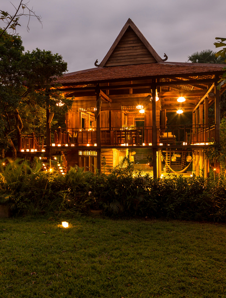 Amansara pavilion illuminated at dusk, warm golden light glowing through wooden shutters onto manicured gardens.
