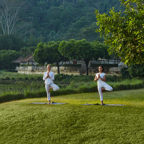 Two people practising yoga on a grass lawn at Amanjiwo, with lush greenery and a building visible across the water beyond.