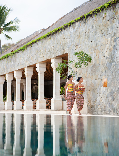 Couple standing by a reflection pool at Amankila, with stone columns and palm trees framing the scene.