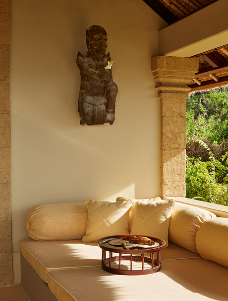 Sunlit spa bath at Amankila with carved wooden mask on wall and natural stone surrounds.