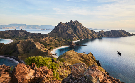 Aerial view of Amandira's rugged coastline with forested peaks meeting calm blue waters and distant mountains.