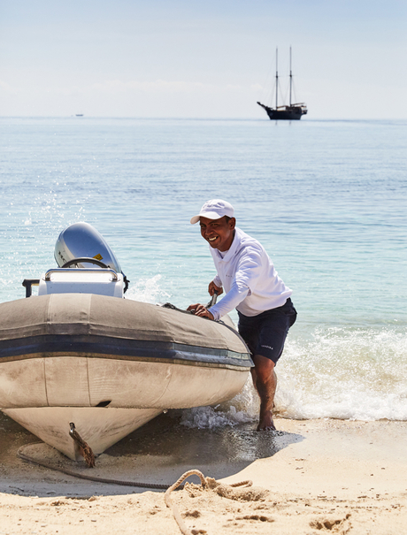 A man in a cap pulls a dinghy ashore at Amandira, Indonesia, with a sailing vessel anchored offshore.